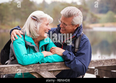 Felice coppia senior appoggiato su di una staccionata in legno a sorridere a ogni altro, vicino Lake District, REGNO UNITO Foto Stock