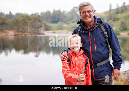 Nonno e nipote insieme in piedi sulla riva di un lago di sorridere alla telecamera, vicino Lake District, REGNO UNITO Foto Stock