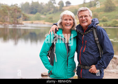 Felice coppia senior in piedi sulla riva di un lago di sorridere alla telecamera, Lake District, REGNO UNITO Foto Stock