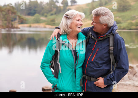 Felice coppia senior in piedi su una riva di un laghetto sorridente ad ogni altro, Lake District, REGNO UNITO Foto Stock