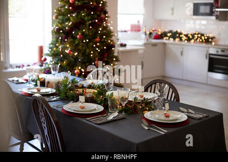 Tavola di natale con impostazione nome ninnolo titolari di carta disposti sulle piastre e verde e rosso e decorazioni con albero di Natale e la cucina in background Foto Stock