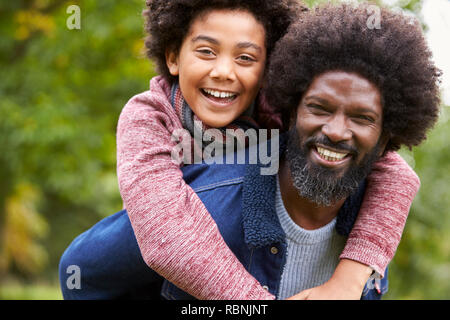 Uomo nero piggybacking sua pre-teen figlio nel parco sia sorridere alla telecamera, close up Foto Stock