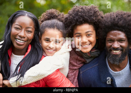 Razza mista famiglia sorridente alla telecamera, genitori piggybacking i bambini nel parco, vicino fino Foto Stock