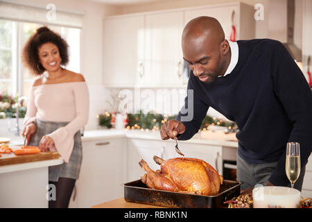 I giovani adulti razza mista giovane preparare la cena di Natale insieme a casa, uomo inumidendo il tacchino arrosto in primo piano, vista frontale, close up Foto Stock