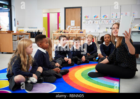 Insegnante femmina tenendo in mano un libro davanti la sua classe della scuola elementare i bambini seduti sul pavimento in un'aula, vista laterale Foto Stock