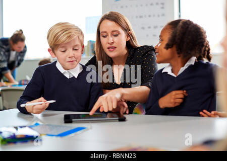 Femmina insegnante di scuola per aiutare due bambini usando un computer tablet al banco in una scuola primaria in aula, vista frontale Foto Stock