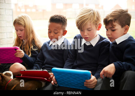 Quattro bambini della scuola primaria di seduta sul pavimento di fronte a una finestra utilizzando i computer tablet durante il tempo di interruzione, close up Foto Stock