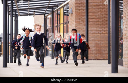 La scuola primaria bambini indossano uniformi di scuola e zaini, in esecuzione su una passerella al di fuori del loro edificio scolastico, vista frontale Foto Stock