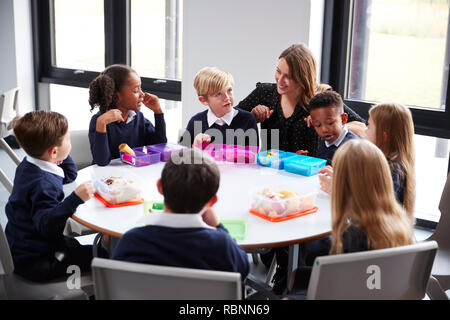 Insegnante femminile inginocchiato a parlare con un gruppo di scuola primaria bambini seduti insieme attorno ad una tavola rotonda di mangiare il loro pranzo al sacco Foto Stock