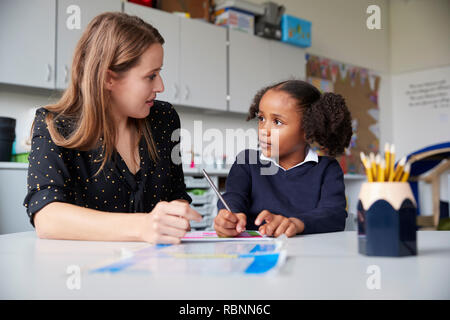 Femmina giovane insegnante di scuola primaria a lavorare uno a uno con un schoolgirl su un tavolo o in un'aula, guardando ogni altro ravvicinato Foto Stock