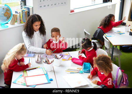 Vista in elevazione della femmina di insegnante di scuola materna seduti ad un tavolo aiutare quattro bambini durante una lezione Foto Stock