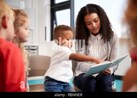 Scuole materne ragazzo puntando in un libro tenuto dall'insegnante femminile, seduta con i ragazzi su sedie in aula, close up Foto Stock