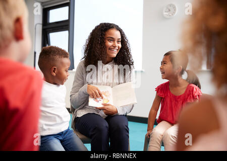 Giovani insegnanti di sesso femminile che mostra una foto in un libro per bambini in una scuola infantile classe seduti su sedie in aula, sulla spalla vista Foto Stock