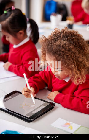 Vista in elevazione del giovane schoolgirl indossano uniformi scolastiche seduto alla scrivania in un infante aula scolastica utilizzando un computer tablet e stilo, close up, verticale Foto Stock