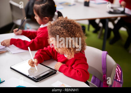Vista in elevazione del giovane schoolgirl indossano uniformi scolastiche seduto alla scrivania in un infante aula scolastica utilizzando un computer tablet e stilo, close up Foto Stock