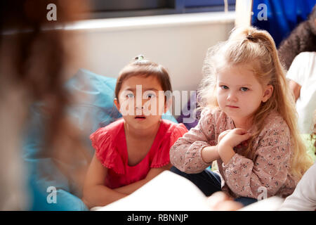 Due scuole materne ragazze seduti sui sacchi di fagioli in un confortevole angolo della classe ascoltando il loro insegnante la lettura di una storia, vista su insegnante di spallamento Foto Stock