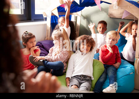 Vista sopra la spalla del bambino insegnante della scuola che mostra un libro a un gruppo di bambini seduti sui sacchi di fagioli in un confortevole angolo della classe, alzando le mani per rispondere a una domanda, il fuoco selettivo close up Foto Stock