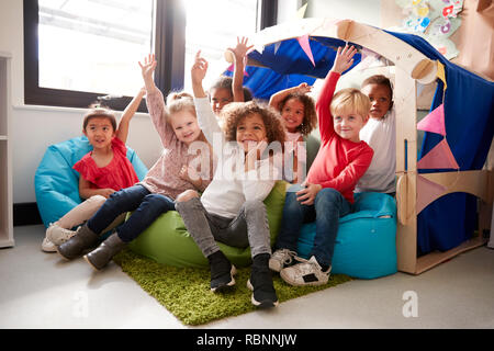 Un multi-etnico gruppo di scuole materne bambini seduti sui sacchi di fagioli in un confortevole angolo della classe, alzando le mani per rispondere a una domanda e a basso angolo, close up Foto Stock