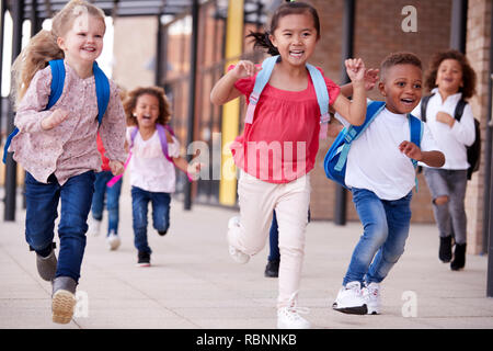 Un gruppo di sorridere multi-etnico i bambini della scuola in esecuzione in una passerella al di fuori delle loro scuole materne edificio dopo una lezione, chiudere fino Foto Stock