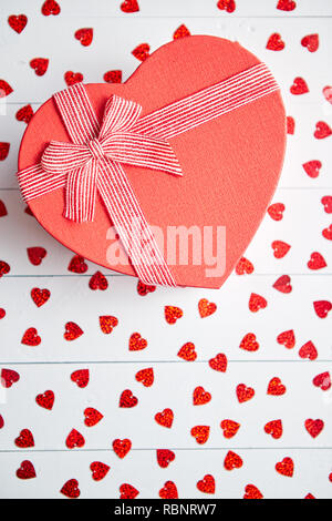 Regalo confezionato posto su a forma di cuore di paillettes rosso su bianco tavolo in legno Foto Stock