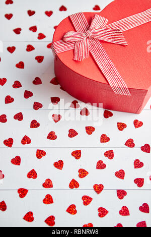 Regalo confezionato posto su a forma di cuore di paillettes rosso su bianco tavolo in legno Foto Stock