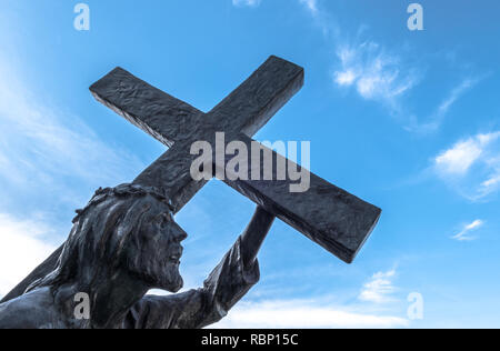Statua di bronzo di Gesù che porta la croce, sulla strada per la sua crocifissione. Ideale per il concetto di pasqua, risurrezione e altri. Cielo blu con il bianco cl Foto Stock