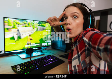 Entusiasti gamer ragazza seduta al tavolo, riproduzione di giochi online su un computer in ambienti chiusi, prendendo un selfie Foto Stock