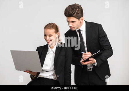 Business sorridente giovane indossando tute isolate su uno sfondo grigio, lavorando insieme con computer portatile, prendendo appunti Foto Stock