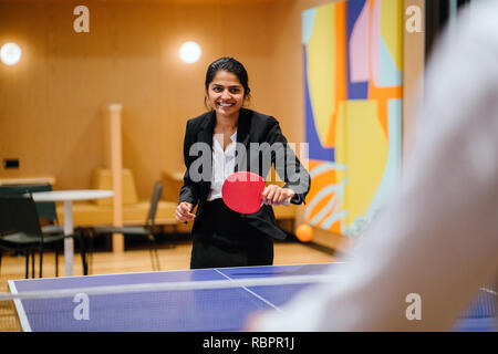 Un giovane indiano donna asiatica in un vestito ride e scherza come gioca a ping pong nel suo ufficio di avvio. Sta avendo un sacco di divertimento con un collega. Foto Stock