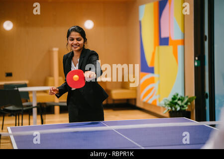 Un giovane indiano donna asiatica in un vestito ride e scherza come gioca a ping pong nel suo ufficio di avvio. Sta avendo un sacco di divertimento con un collega. Foto Stock