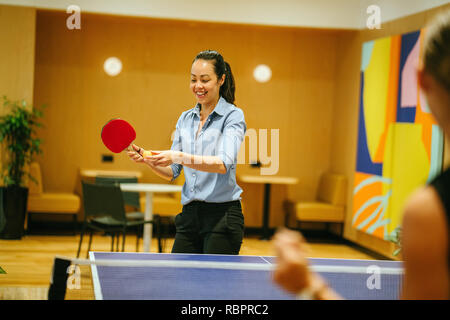 Un attraente donna asiatica gioca a ping-pong con un collega in un elegante spazio coworking ufficio dove lavora all'avvio. Lei è sorridente. Foto Stock