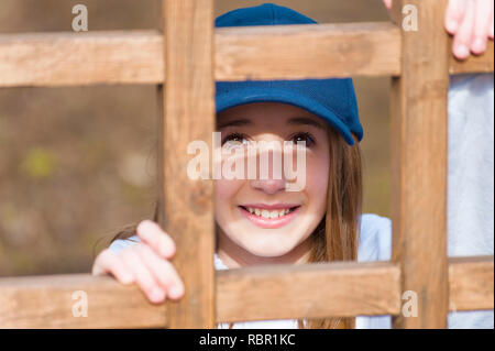 Indossando un tappo a sfera e un grande sorriso di una giovane ragazza guarda la fotocamera da behine un quadrato in legno a traliccio. Foto Stock