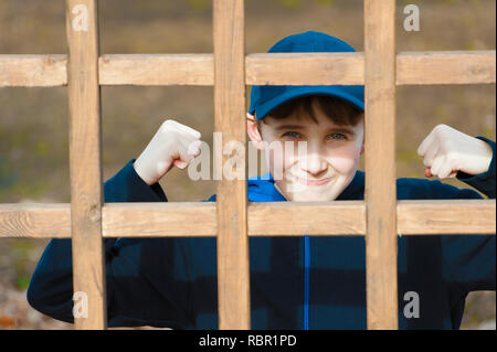 Un giovane ragazzo solleva si adatta con un sorriso a tenuta in posa per la telecamera dietro un reticolo di legno. Foto Stock