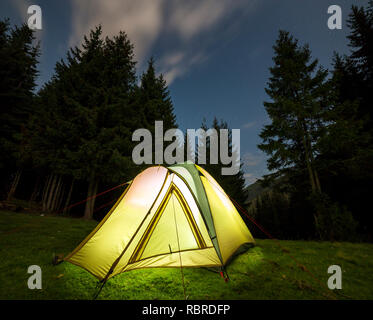 Gli escursionisti turistici tenda illuminata dall'interno sul verde foresta erbosa cancellazione tra alti pini sotto blu scuro cielo stellato sulla montagna lontano indietro Foto Stock