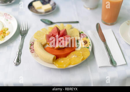 Una piastra su un tavolo con tropicale naturale di frutta, arancio, guaiava, banana e ananas. Che circonda la piastra ci sono una forchetta, un coltello e un cucchiaino. Foto Stock