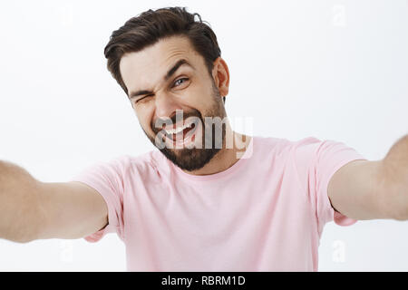 Studio shot del carismatico felice ed entusiasta di bello macho uomo con barba e capelli scuri winking e sorridente con gioia le palme di trazione verso la telecamera tenendo lo smartphone e tenendo selfie Foto Stock