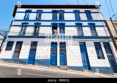 Blu e bianco blocco alloggiamento su una ripida collina posizione, da una strada, a Valparaiso, Cile, con uno dei residenti la lettura di una carta. Foto Stock