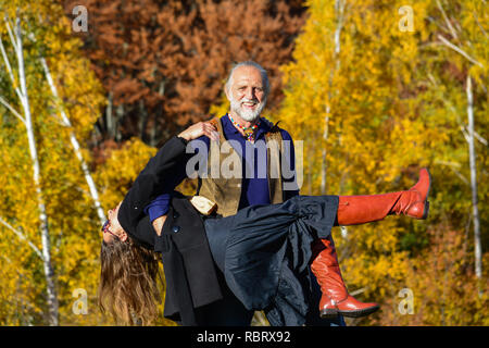 Felice, vitale coppia di anziani ballare e divertirsi in un luminoso giorno di sole sul prato di montagna e bosco di betulle, in background Foto Stock