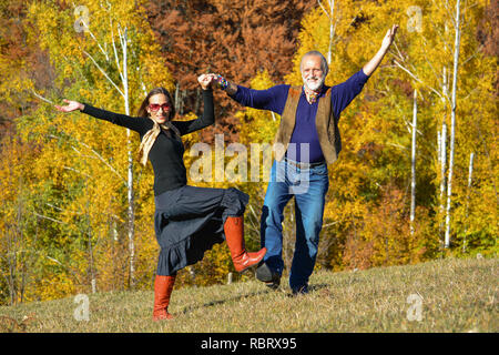 Felice, vitale coppia di anziani per godersi la vita, ballare e divertirsi in un luminoso giorno di sole sul prato di montagna e bosco di betulle, in background Foto Stock