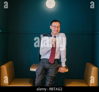 Ritratto di un uomo d'affari asiatici in un vestito e un tirante appoggiata contro un elegante stand blu nel suo ufficio. Egli sorride con fiducia. Foto Stock