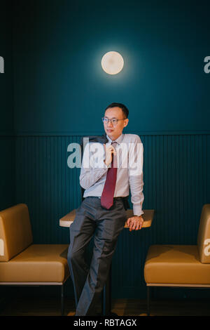 Ritratto di un uomo d'affari asiatici o professional (avvocato, banchiere, etc) appoggiata contro un tavolo nel suo ufficio. Egli è alto e sottile e fiducioso. Foto Stock