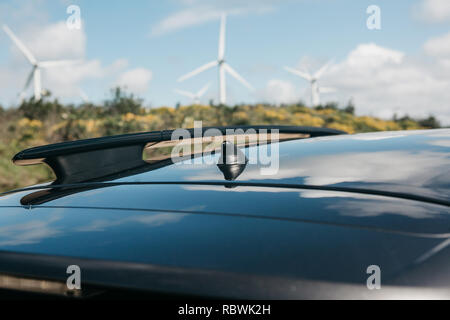 Foto concettuale con una prospettiva alternativa con il fuoco selettivo sulla antenna sul tetto della vettura e sfocata mulini a vento o le turbine eoliche in background. Foto Stock