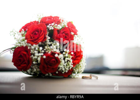 Close-up di una coppia di anelli di nozze, dietro di loro un mazzo di fiori di colore rosso. Foto Stock