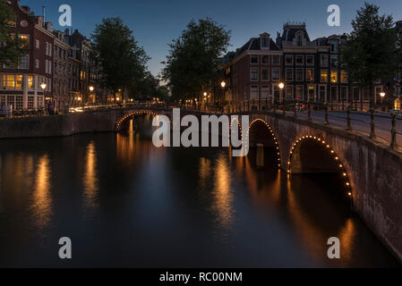 Canale di Amsterdam e il bridge al tramonto. Il canale è calma e le luci intorno i ponti arcate sono riflettendo sull'acqua liscia. Foto Stock