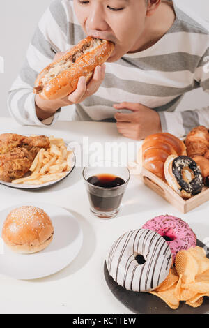 Ritratto di uomo asiatico seduto e snacking ciambelle isolate su sfondo bianco. Uomo grasso concetto di dieta Foto Stock