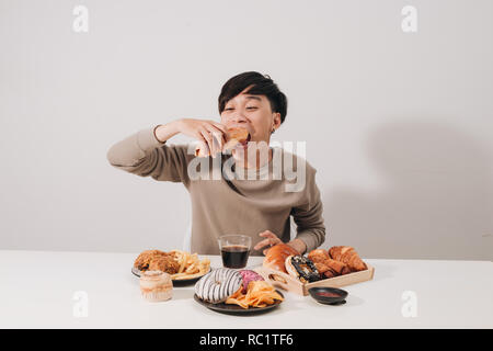 Ritratto di uomo asiatico seduto e snacking ciambelle isolate su sfondo bianco. Uomo grasso concetto di dieta Foto Stock