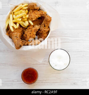 Gustoso un fast food: pollo fritto gambe, ali piccante, patatine fritte e strisce di gara nella scatola di carta, salsa e birra fredda bianco su una superficie di legno, vista dall'alto. Foto Stock