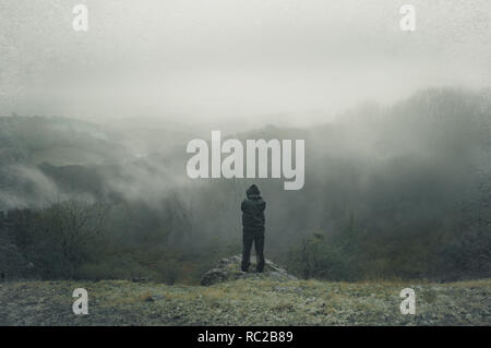 Un lone incappucciati figura sulla cima di una collina che si affaccia su tutta la campagna su una nebbia, inverni piovosi giorno. Con una deliberata grunge, sgranate, disattivato modifica. Foto Stock