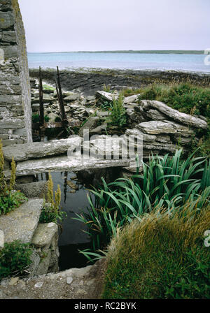 Hookin mulino ad acqua, Papa Westray, Orkney. Rovinato undershot corn mill accanto alla spiaggia con resti di canale di acqua e ferro quadro della ruota ad acqua Foto Stock