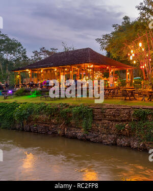 Serata di scena illuminata con un ristorante esterno dal fiume nella giungla tropicale, Pai, Thailandia Foto Stock